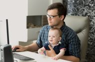 Homem trabalha no computador com bebê no colo. Bebê sorridente olha para frente.