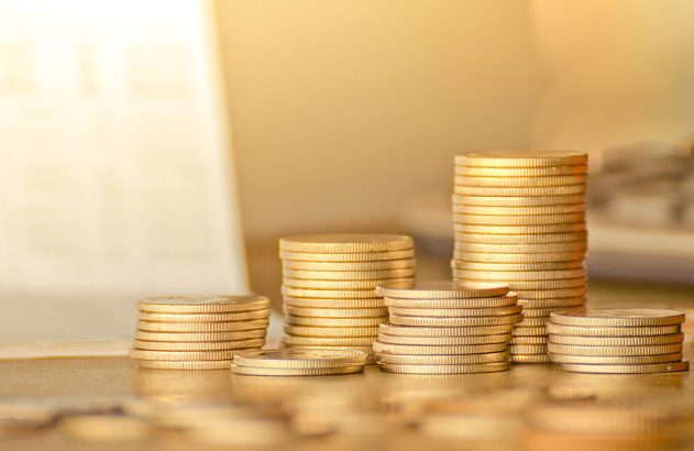 Pilhas de moedas douradas de tamanhos variados sobre uma superfície, com fundo desfocado e iluminado.