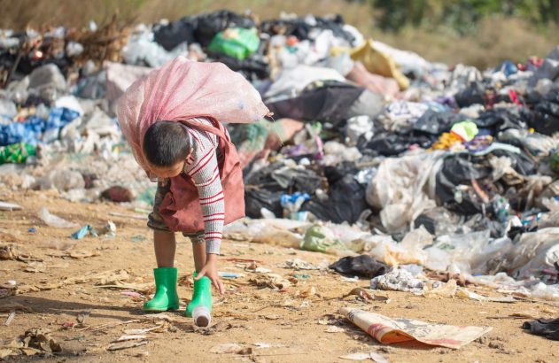 Criança catando garrafa plástica em um lixão com um saco nas costas.