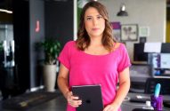 Mulher em pé, camisa rosa, segurando tablet em ambiente de escritório.