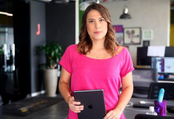 Mulher em pé, camisa rosa, segurando tablet em ambiente de escritório.