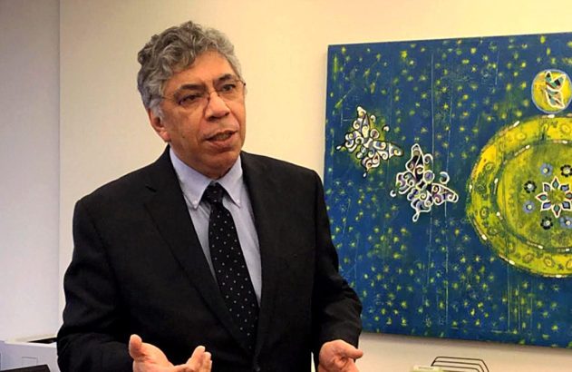 Homem de terno gesticulando enquanto fala, com um quadro de arte colorido ao fundo.