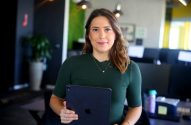 Mulher de camisa verde segura tablet, sorrindo para a câmera em um escritório.