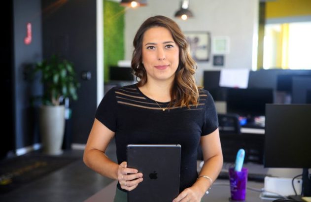Mulher em pé em um escritório, segurando um tablet e olhando para a câmera.