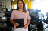 Mulher com blusa rosa e calça branca, segurando um tablet, em pé num escritório com mesas e computadores.