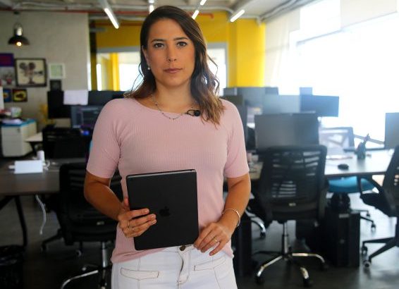 Mulher com blusa rosa e calça branca, segurando um tablet, em pé num escritório com mesas e computadores.