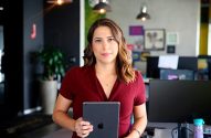 Mulher de vestido vinho segura tablet em escritório, olhando para frente.