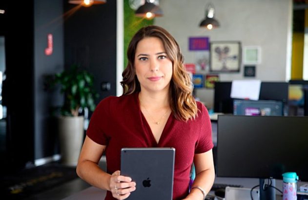 Mulher de vestido vinho segura tablet em escritório, olhando para frente.