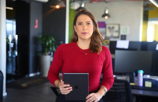 Mulher de suéter vermelho segurando tablet e olhando para a câmera em escritório.