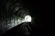 Um túnel ferroviário escuro com trilhos curvando-se em direção a uma luz intensa na saída.