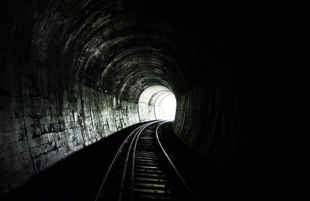 Um túnel ferroviário escuro com trilhos curvando-se em direção a uma luz intensa na saída.