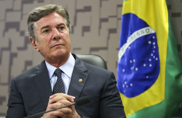 Homem de terno e gravata, mãos unidas, sentado. Bandeira do Brasil ao lado, em ambiente formal.