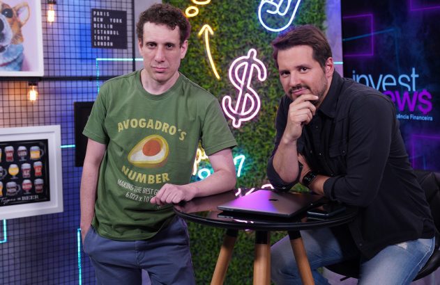 Dois homens posam em estúdio de TV. Um em pé, outro sentado com laptop. Fundo com símbolos de dólar e logo Invest News.