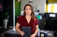 Mulher em pé num escritório, segurando um tablet e olhando para a câmera.