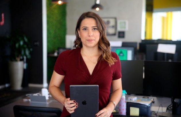 Mulher em pé num escritório, segurando um tablet e olhando para a câmera.