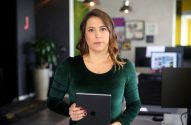 Mulher de blusa verde em pé segura um tablet, olhando para a câmera em um escritório.