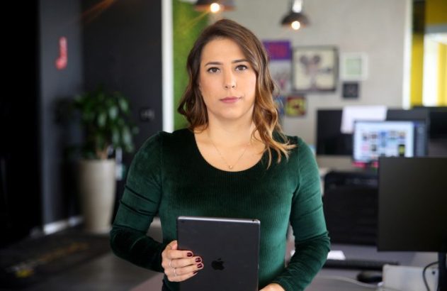 Mulher de blusa verde em pé segura um tablet, olhando para a câmera em um escritório.