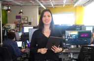 Mulher sorrindo, segurando tablet, em escritório com pessoas trabalhando.