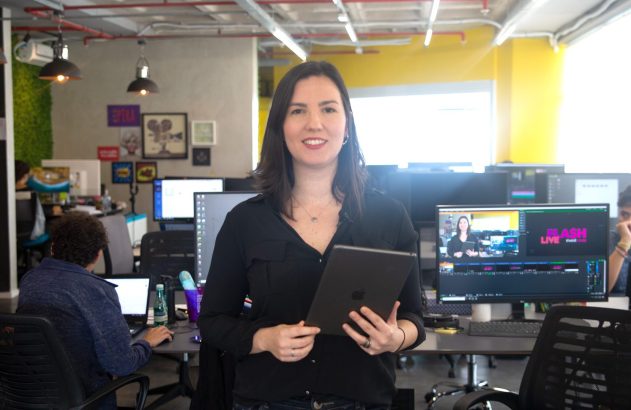 Mulher sorrindo, segurando tablet, em escritório com pessoas trabalhando.