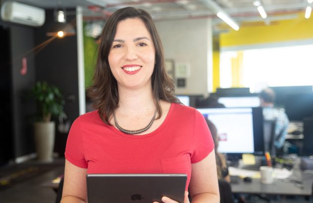 Mulher sorrindo, em camiseta vermelha, segura um tablet. Fundo de escritório com pessoas e telas desfocadas.