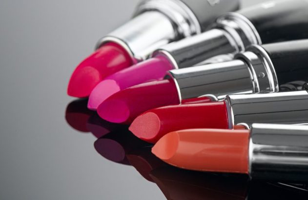 Fileira de batons coloridos (rosa, vermelho, laranja) e em escala de cinza, dispostos em superfície reflexiva.