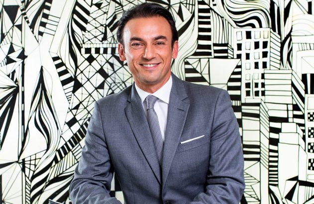 Homem sorrindo de terno cinza e gravata, olhando para a câmera. Fundo com padrões preto e branco.