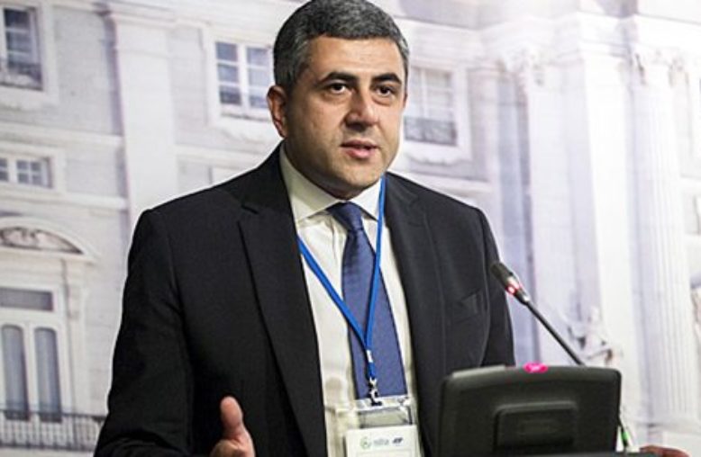 Homem de terno gesticula e fala em evento, com microfone e tela à sua frente.