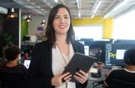 Mulher sorridente de blazer e camisa branca, segurando um tablet, em um escritório moderno.