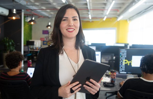 Mulher sorridente de blazer e camisa branca, segurando um tablet, em um escritório moderno.