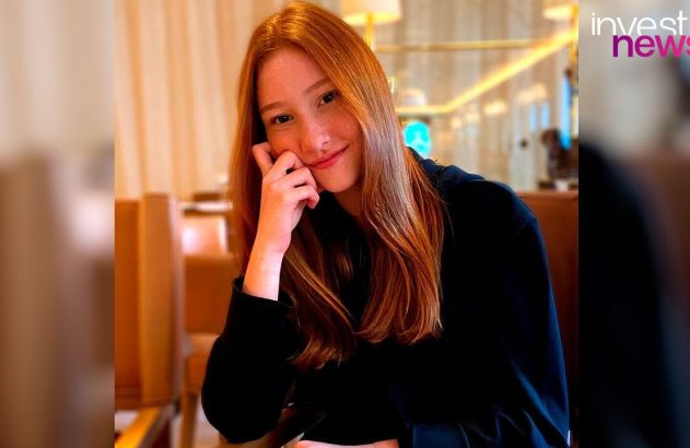 Jovem mulher ruiva sorrindo, com a mão no rosto, usando blusa preta. Fundo desfocado de ambiente interno.