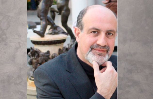 Homem com barba grisalha e roupa escura, tocando o queixo. Estátuas de bronze ao fundo.