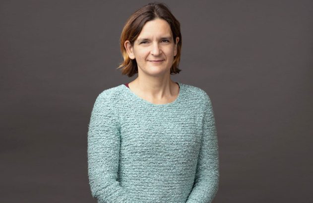 Retrato de mulher com cabelo castanho curto, blusa azul-clara, sorrindo levemente para a câmera, fundo cinza liso.