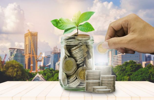 Mão colocando moeda em pote com moedas e broto. Pilhas de moedas e cidade no fundo. Símbolo de investimento.