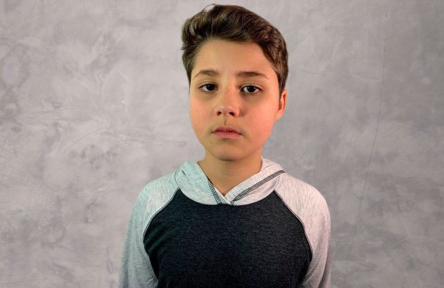 Menino jovem, com lágrimas no rosto e capuz cinza, olhando sério para a câmera.