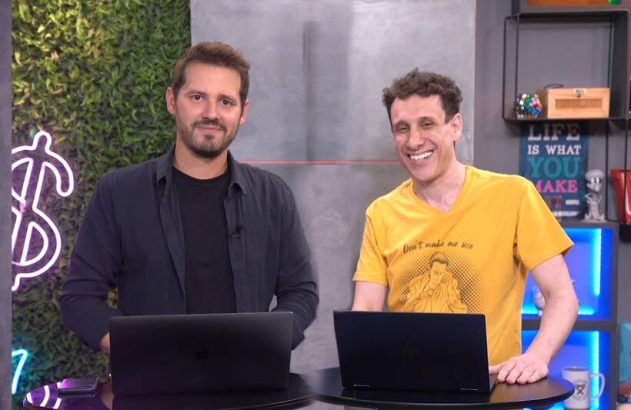 Dois homens sorridentes em pé, atrás de laptops em um estúdio. Um sorri abertamente, outro calmamente.