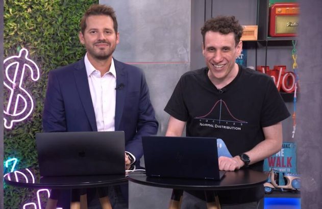 Dois homens sorrindo em estúdio com laptops. Um de terno, o outro com camiseta. Fundo com letreiro neon de cifrão.