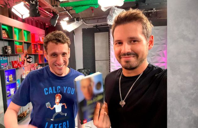 Dois homens sorrindo em estúdio. O da direita segura um livro e o da esquerda veste uma camiseta "CALC YOU LATER!".