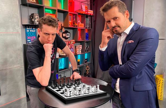 Dois homens em um ambiente moderno. Um joga xadrez pensativo, o outro posa com a mão no queixo.