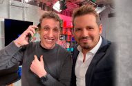 Um homem sorridente atende o telefone e aponta para si, ao lado de outro homem sorrindo em um estúdio.