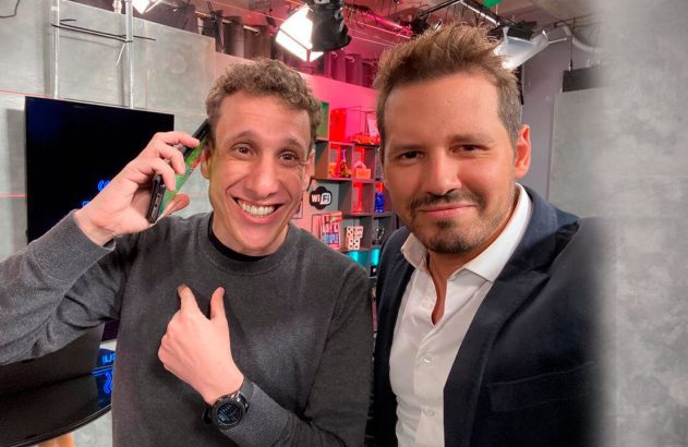 Um homem sorridente atende o telefone e aponta para si, ao lado de outro homem sorrindo em um estúdio.