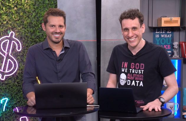 Dois apresentadores sorrindo em um estúdio de notícias com laptops.