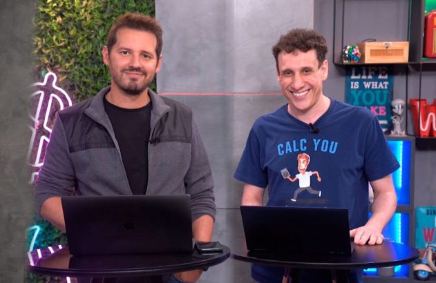 Dois homens sorrindo, um de camiseta azul "CALC YOU", em pé atrás de laptops em um estúdio.