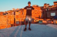 Jovem com camiseta "Favelado & Investidor" de braços abertos em telhado de favela ao pôr do sol.