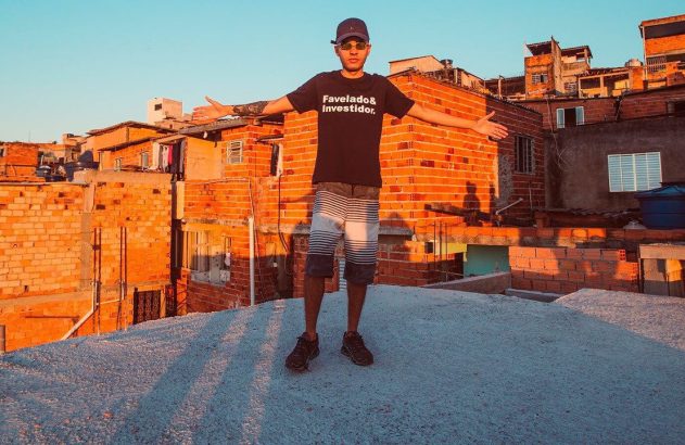 Jovem com camiseta "Favelado & Investidor" de braços abertos em telhado de favela ao pôr do sol.
