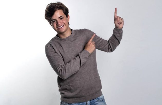 Jovem sorridente, com moletom e jeans, aponta com dois dedos para o lado e para cima.