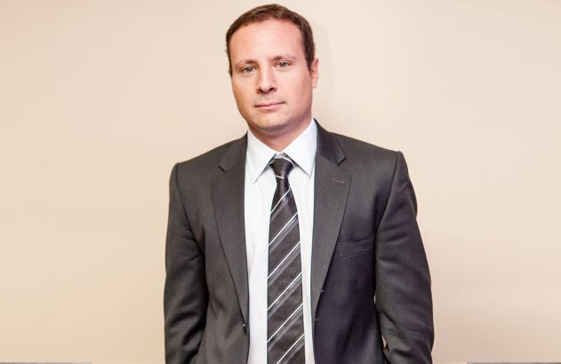 Homem de terno cinza, camisa branca e gravata listrada, olhando para a câmera.