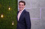 Homem sorridente de terno azul e camisa rosa em pé, contra fundo de parede verde com luzes decorativas.