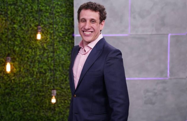 Homem sorridente de terno azul e camisa rosa em pé, contra fundo de parede verde com luzes decorativas.