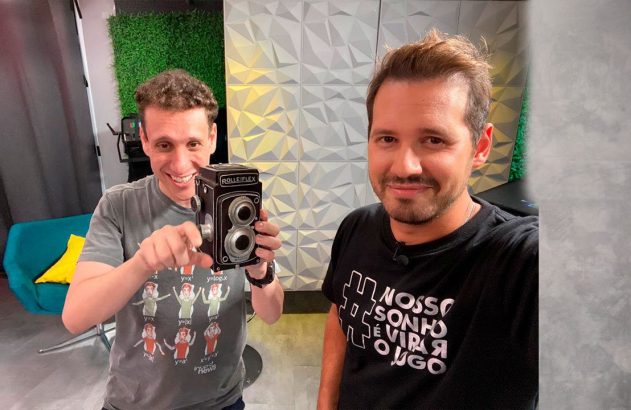Dois homens sorrindo: um segura câmera Rolleiflex e aponta, o outro posa para selfie.