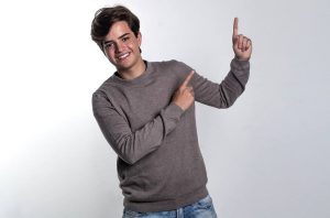 Jovem sorridente, com moletom e jeans, aponta com dois dedos para o lado e para cima.